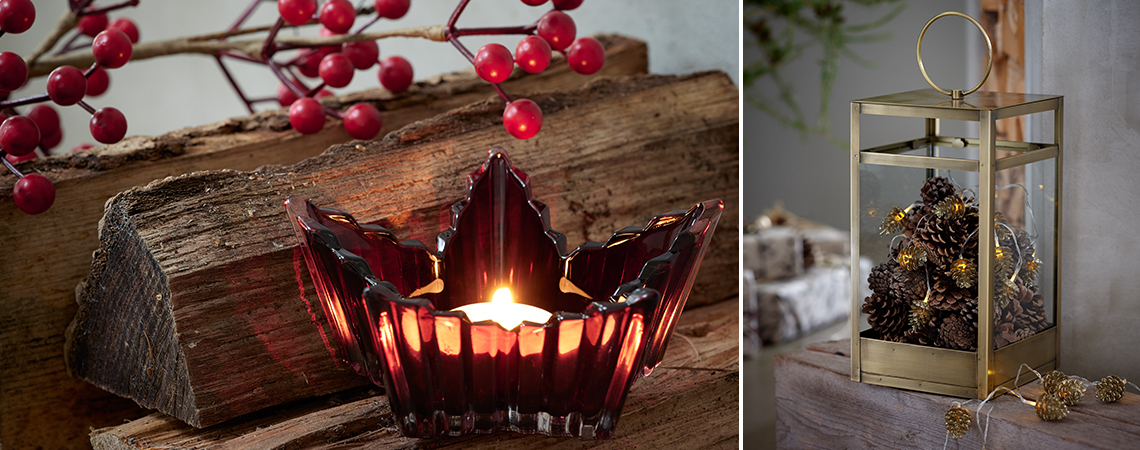 Tea light holder in glass in front of logs and branches with berries and a golden lantern with string lights inside