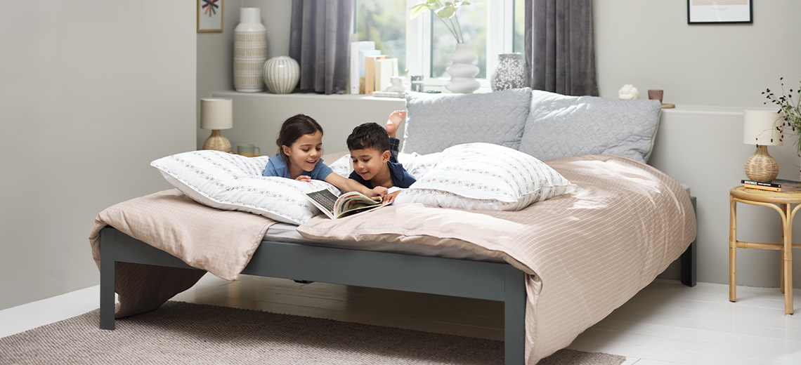 Two children reading a book on double bed with duvet and duvet cover, pillows, and cushions