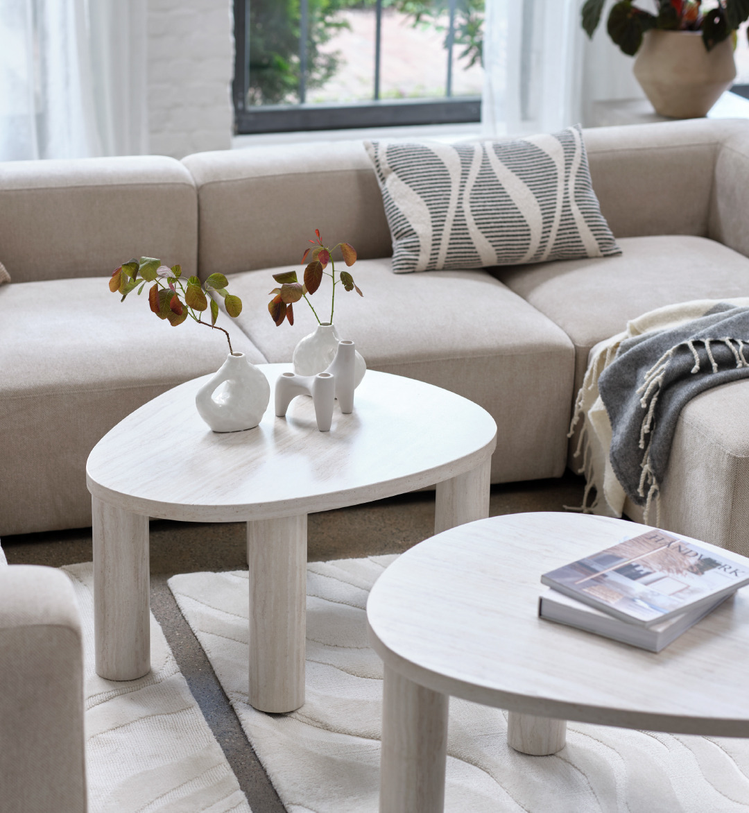 A beige and black decorative cushion on top of a sand coloured modular sofa and two white marble coloured coffee tables