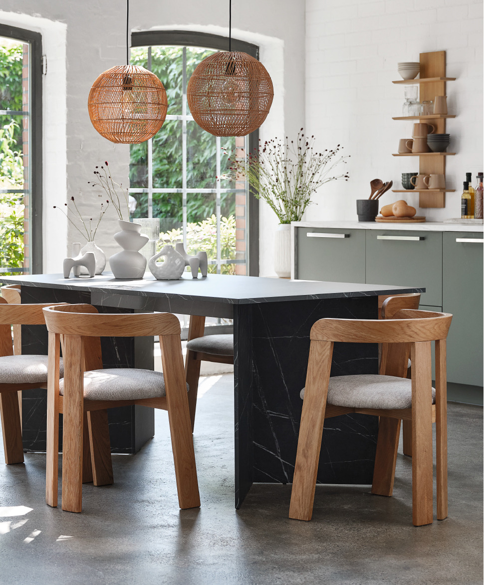 A black marble dining table five oak and beige dining chairs below two rattan light pendants and a wooden wall shelf with ceramic mugs and bowls