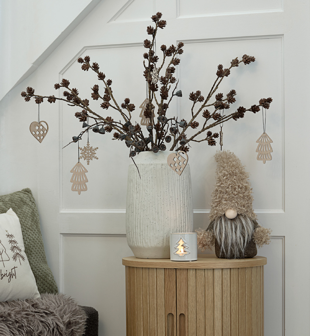 An oak end table with a vase and plant decorated with Christmas ornaments, placed next to a Christmas candle and elf