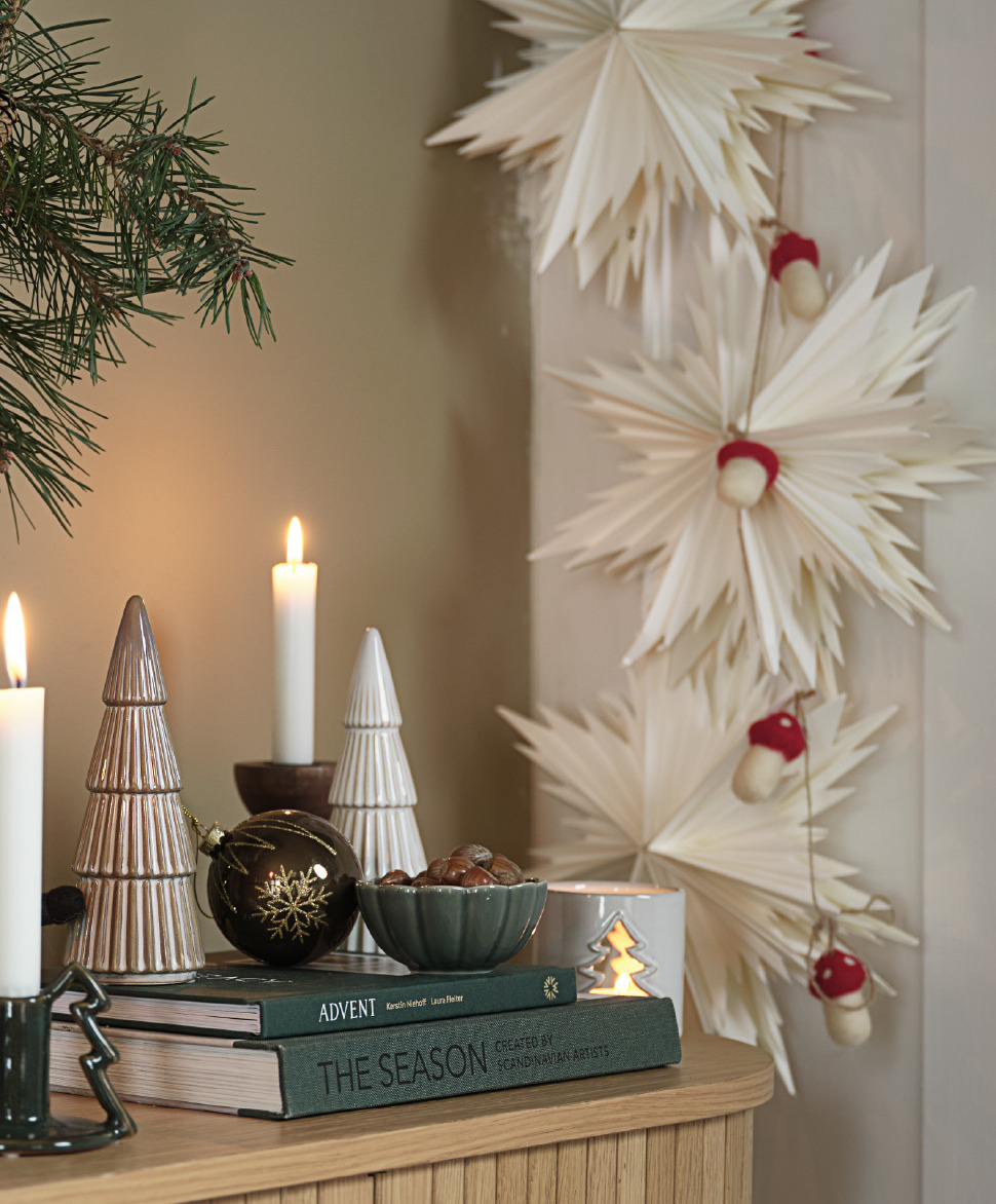 White Christmas stars hanging off a wall next to ceramic Christmas tree decorations on a sideboard