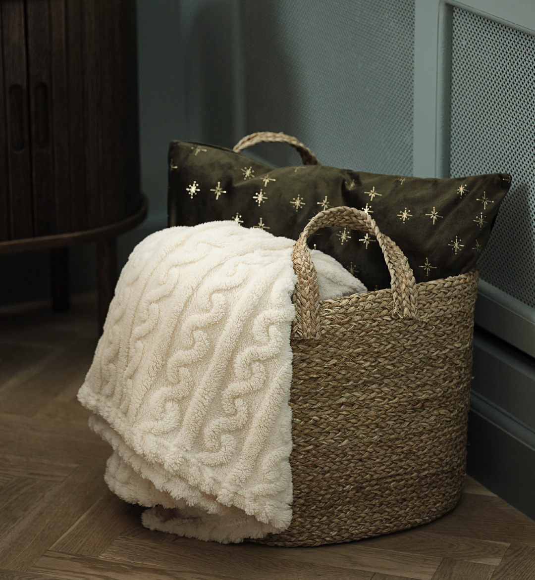 A green decorative cushion and white throw inside of a storage basket