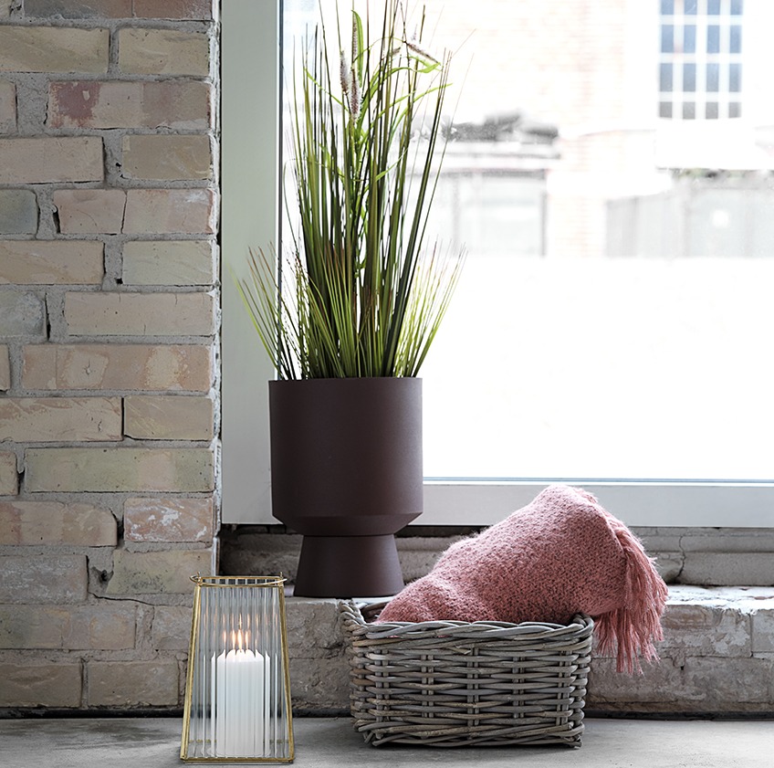 Plant pot with artificial plant, lantern, and basket with a pink throw in front of a window 