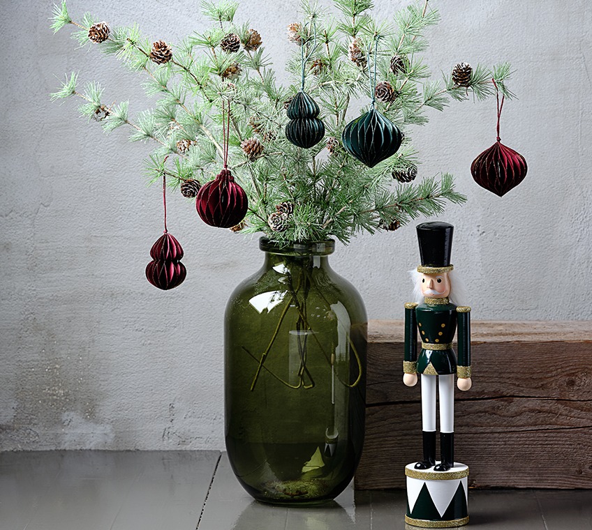 FELIX vase filled with BURR artificial twigs, decorated with PYROP ornaments in green and red. Beside it EUKLAS figurine Green glass vase filled with artificial twigs with pinecones, decorated with Christmas ornaments in green and red. Beside it a Christmas figurine