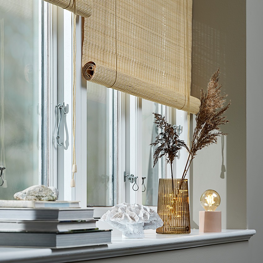 BYRE roller blinds, EJLEV plant pot, TORLEK ornament, ROY vase, JENS scented candle and ARNOLD battery lamp Bamboo roller blinds in a window with plant pot, ornament, vase, scented candle and battery lamp in the windowsill